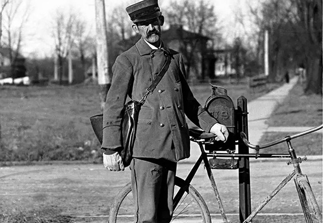 A black-and-white photograph of a postal carrier standing next to his bicycle and a customer's mailbox.
